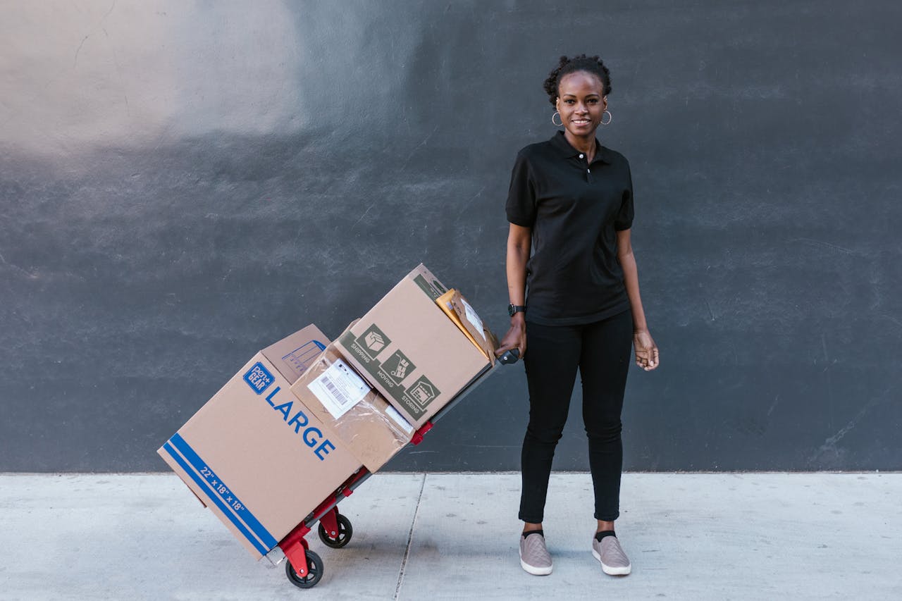 services-03 Black woman in uniform pulling a dolly with large boxes, photographed outdoors against a dark wall.